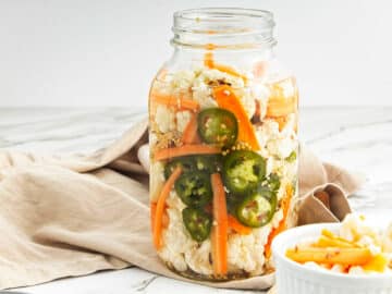 Pickled vegetables in jar with beige cloth behind and small white bowl of vegetables to the right.