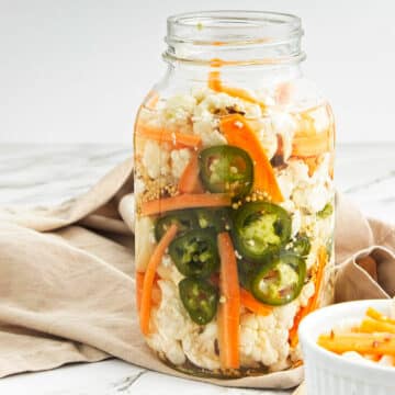 Pickled vegetables in jar with beige cloth behind and small white bowl of vegetables to the right.