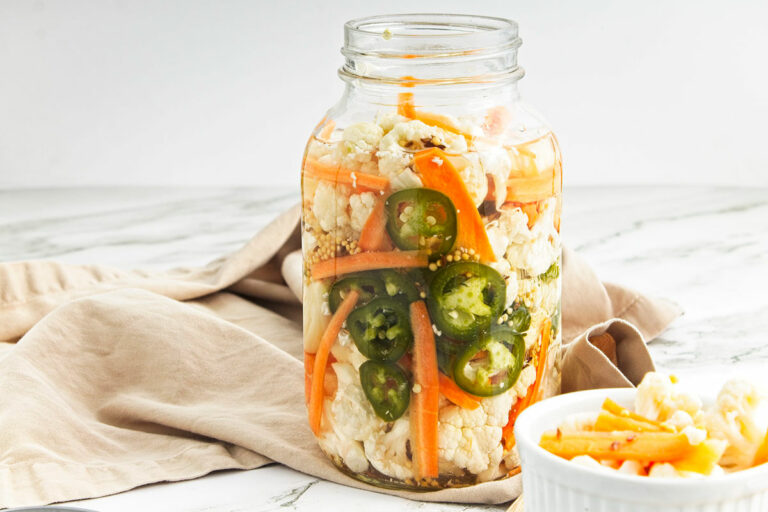Pickled vegetables in jar with beige cloth behind and small white bowl of vegetables to the right.