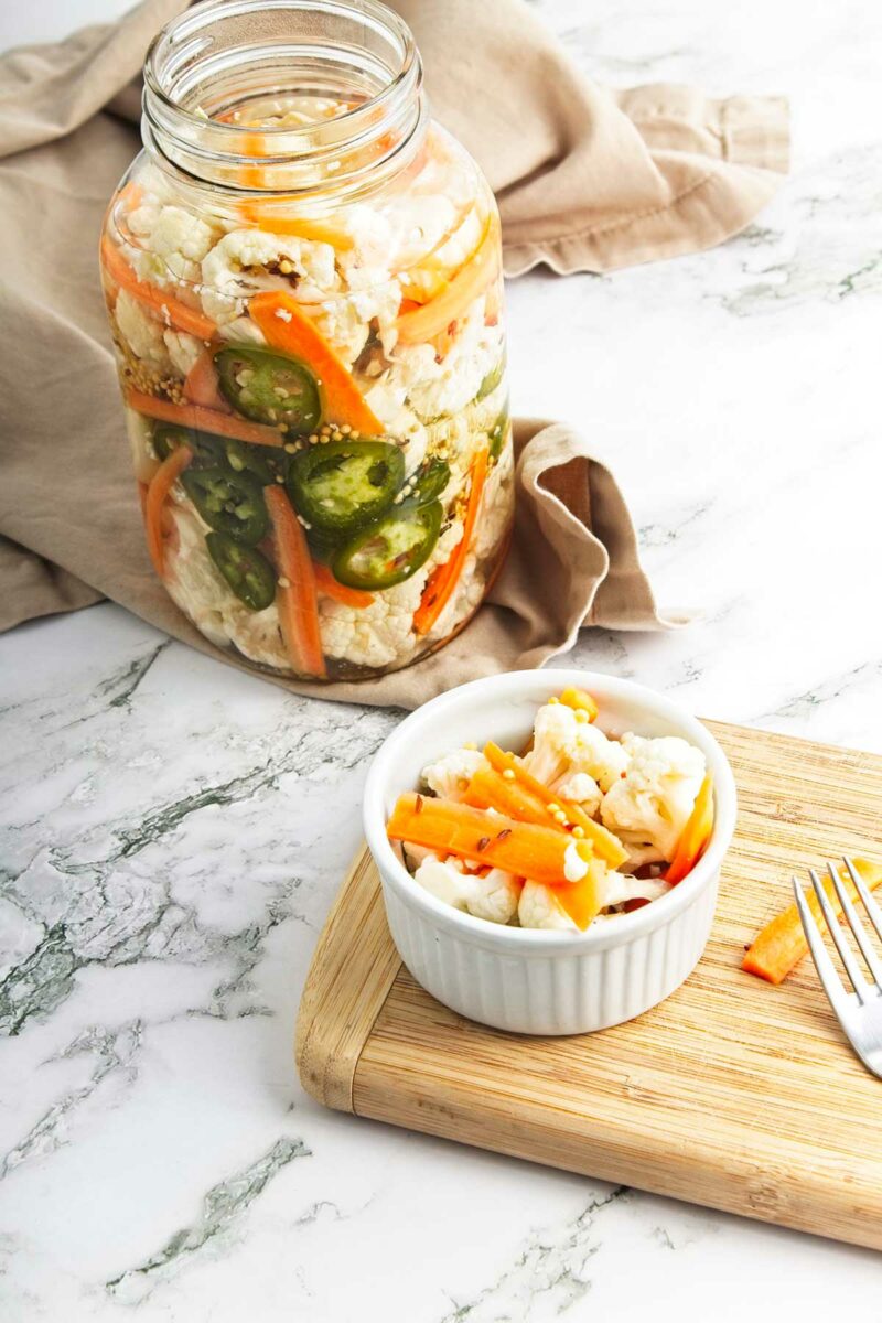 Pickled vegetables in jar with small white bowl on chopping board with fork in front.