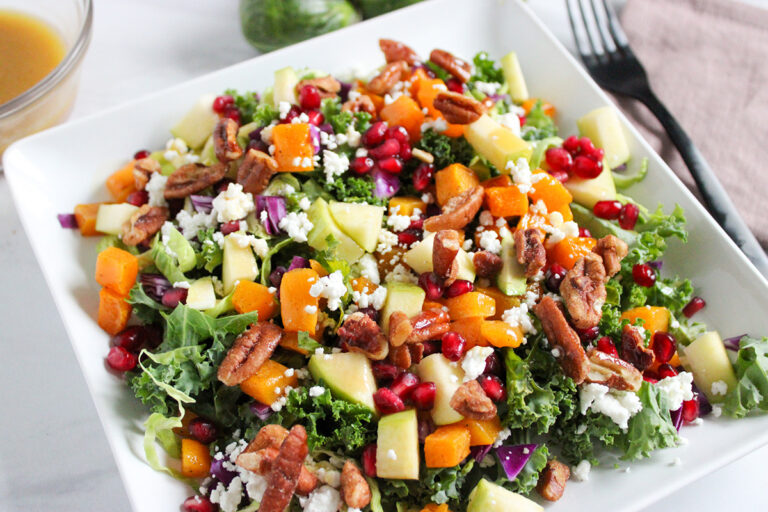 Kale and pomegranate salad in white square dish with fork to the right and dressing behind.