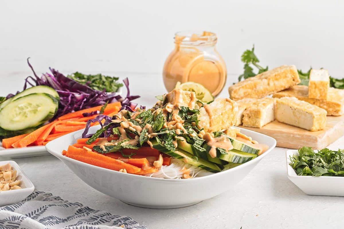 Peanut Tofu Noodles in bowl with veggies and tofu behind and jar of peanut sauce.